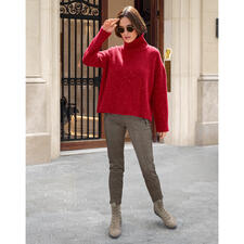 Beige Frau in rotem Pullover und karierten Schlupfhosen steht vor einer Gebäudefassade.