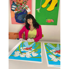 Anna Rupprecht Frau mit schwarzem Haar in pinkem Blazer sitzt an Tisch und signiert Kunstwerke.