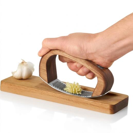 Hand hält AdHoc Knoblauchwippe über Holzbrett mit einer Knoblauchzehe.