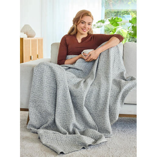 Ideal auch als leichte Decke für gemütliche Stunden auf der Couch. Frau sitzt auf einem Sofa mit einer jacquard Decke umhüllt.