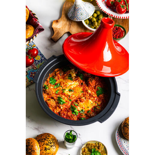 Tajine Peugeot Saveurs mit rotem Deckel, gefüllt mit buntem Gemüse und Kräutern, auf einem Tisch mit Beilagen.