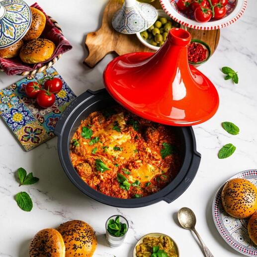 Tajine Peugeot Saveurs mit rotem Deckel, gefüllt mit buntem Gemüse und Kräutern, auf einem Tisch mit Beilagen.