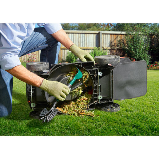 Person reinigt einen umgedrehten Rasenmäher mit einem Werkzeug, während Grasreste entfernt werden.