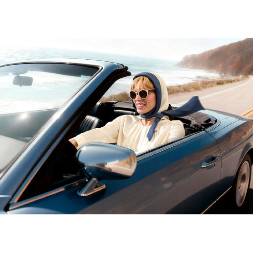 Frau in einem Cabrio mit Kopftuch und Sonnenbrille fährt an Küste entlang.