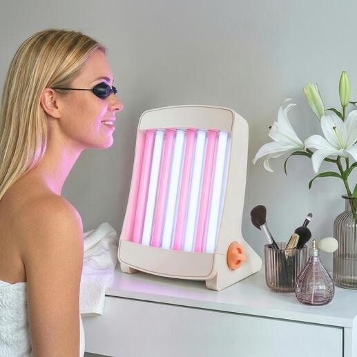 Gesichtsbräuner mit pinken und blauen Lampen, steht auf einem Tisch neben Kosmetikartikeln.