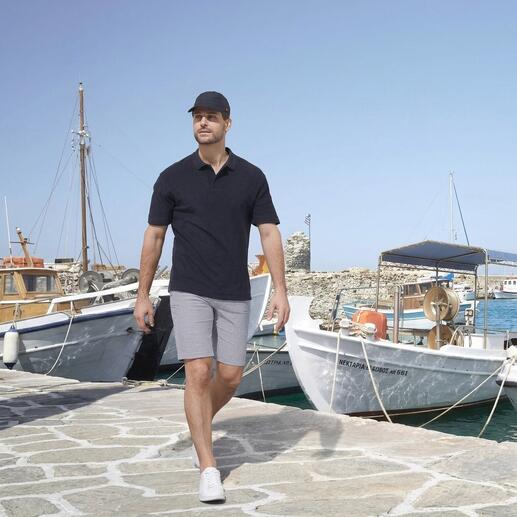 Mann läuft an einem Hafen in kurzen hellgrauen Bermudas und schwarzem Polo mit Booten im Hintergrund.
