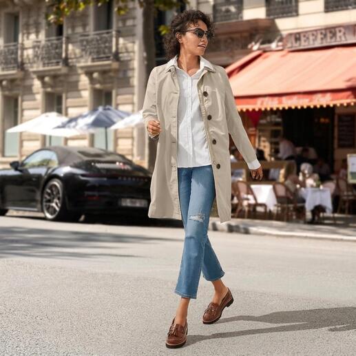 Frau in beigem Trenchcoat und weißen Sneakers, die durch eine belebte Straße mit Cafés geht.