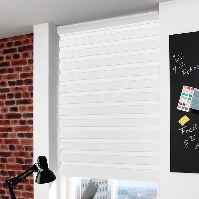 Ein kurzer Zug an der Kette – und schon können Sie Ihr Rollo von blickdicht auf halb oder ganz geöffnet einstellen. Weißes Rollo, das an einem modernen Fenster montiert ist, im Hintergrund sind Ziegelmauern sichtbar.