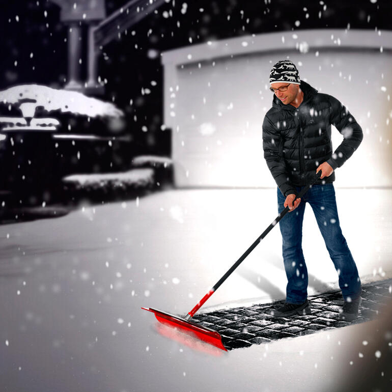 Mann schaufelt Schnee mit Schneeade® vor einer Garage in einer verschneiten Umgebung.