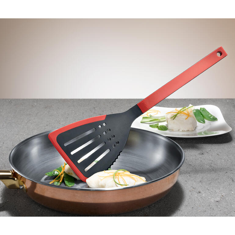 Mit der gezahnten Seite lassen sich Zutaten und Gerichte bereits in der Pfanne zerkleinern. Pfannenwender „Flip it“ liegt neben einem Fischfilet in einer Pfanne.