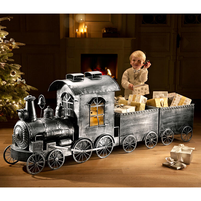 Nostalgie-Zug aus Metall mit Geschenken, im Hintergrund ein dekorierter Weihnachtsbaum.