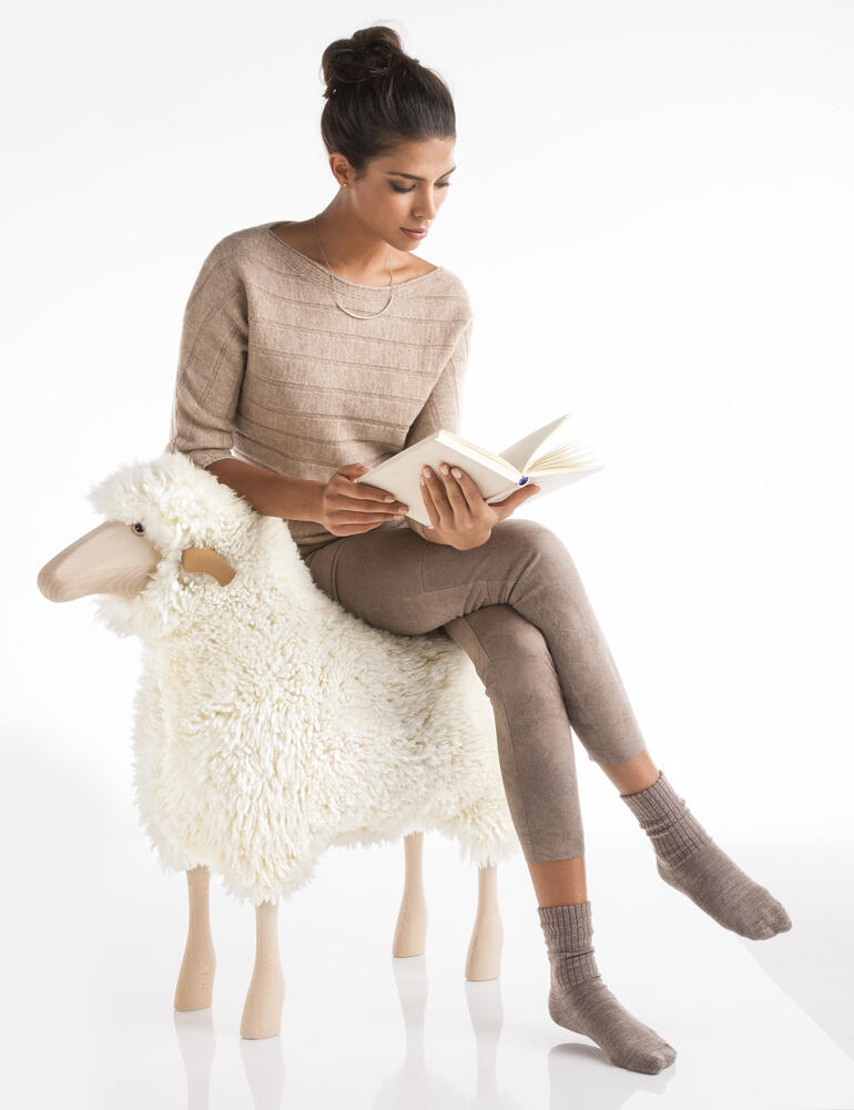 Die Massivolz-Unterkonstuktion ist extrem belastbar, so dass Sie das Designobjekt auch als Hocker nutzen können. Frau sitzt auf einem Schaf und liest ein Buch.