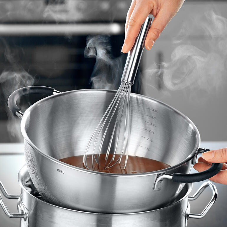 Über dem Wasserbad eignet sich die Schüssel hervorragend zum Schmelzen von Schokolade. Hand mit einem Schneebesen in einer dampfenden Rührschüssel über dem Wasserbad.