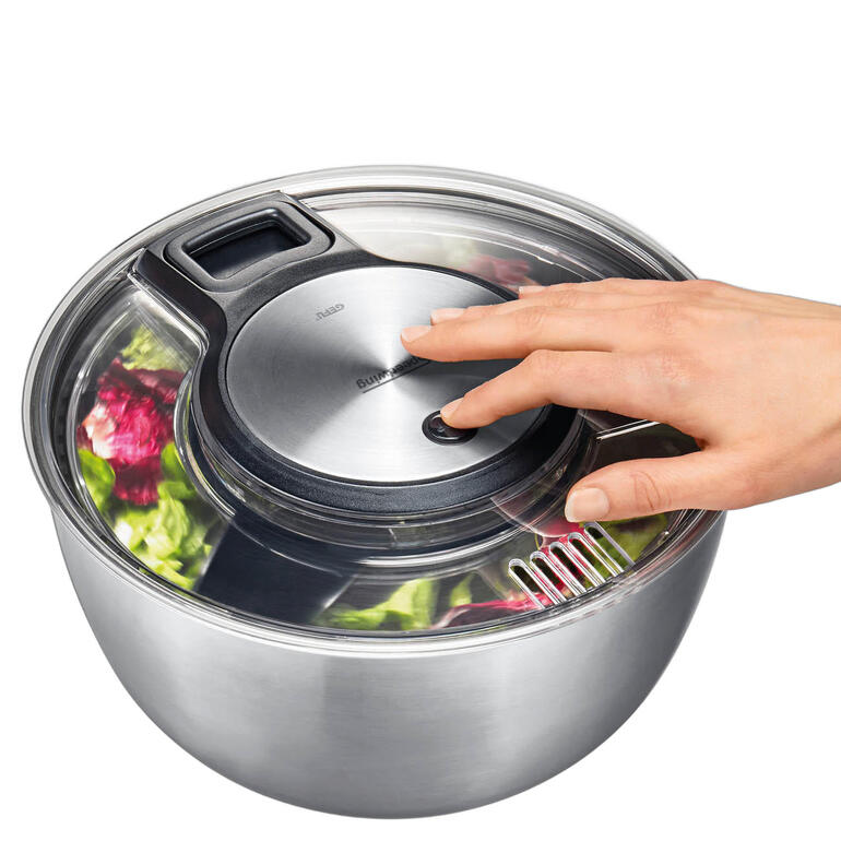 Salatschleuder auf einem Tisch mit frischem Salat und Besteck.