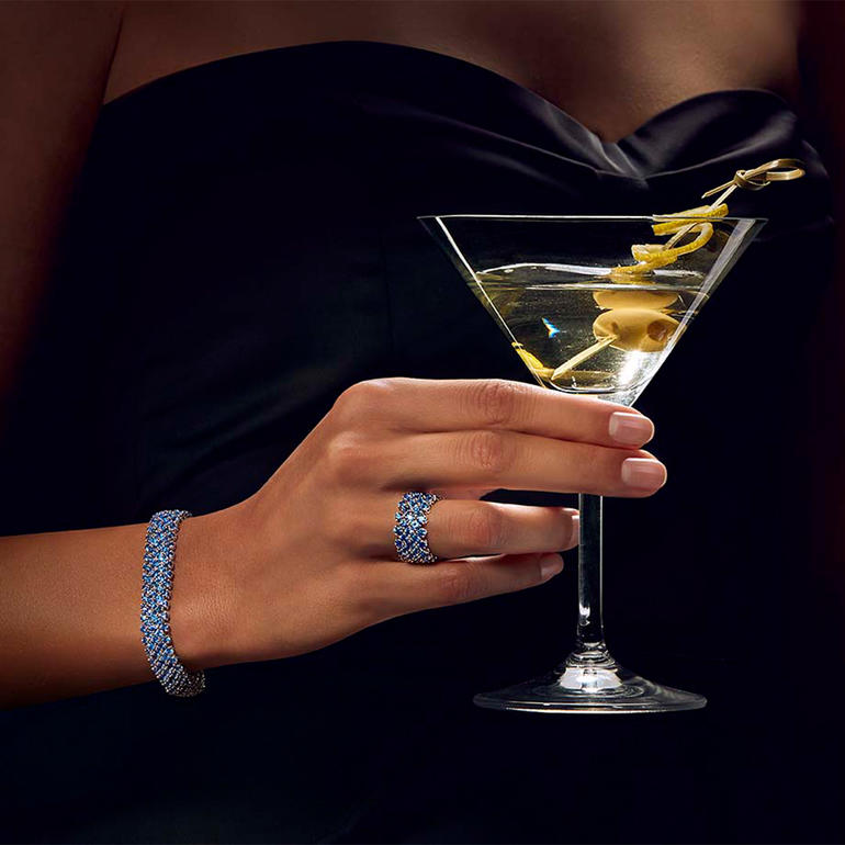 Hand mit einem blauen Saphirring, die einen Cocktail hält.