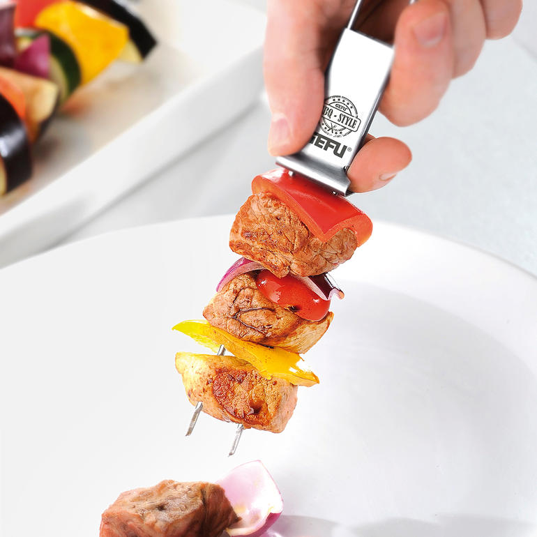 Einfach die Abstreifhilfe nach vorn schieben und Ihr Grillgut gleitet sicher auf den Teller. Hand mit einem Doppel-Grillspieß, der mit Gemüse und Fleisch belegt ist