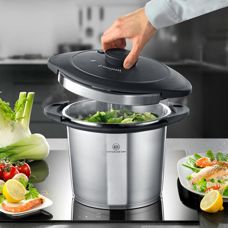 Silberner Schnellkochtopf mit schwarzem Deckel, der gerade geöffnet wird, mit Gemüse im Topf.
