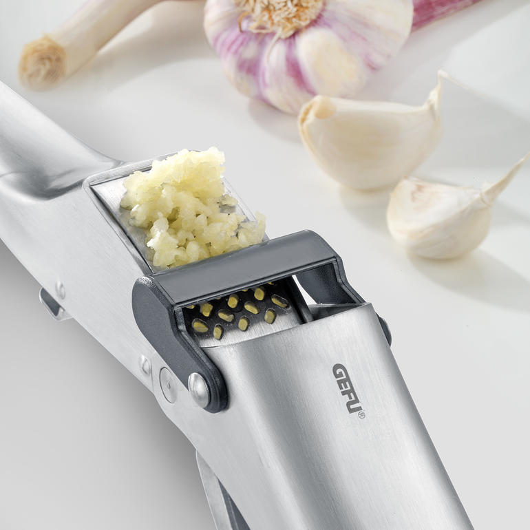 Knoblauchpresse Garlico Knoblauchpresse mit zerdrücktem Knoblauch neben ganzen Zehen