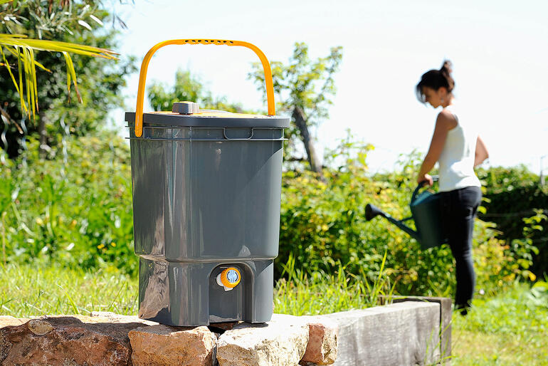 BioMix Flüssigdünger-Bereiter in einem Garten, im Hintergrund arbeitende Frau.