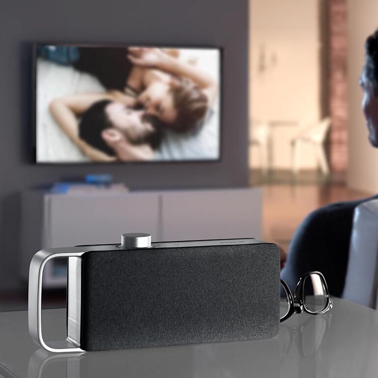TV-Hörverstärker OSKAR, auf einem Tisch stehend, mit einem Fernseher im Hintergrund.
