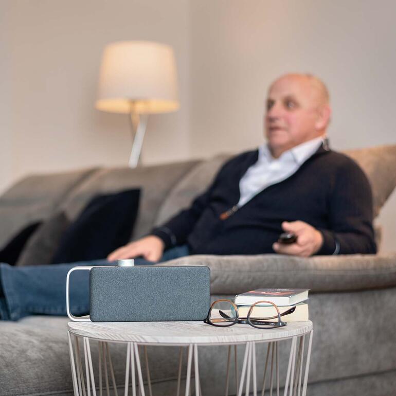 Älterer Mann sitzt auf einem Sofa, schaut fern und nutzt den TV-Hörverstärker OSKAR.