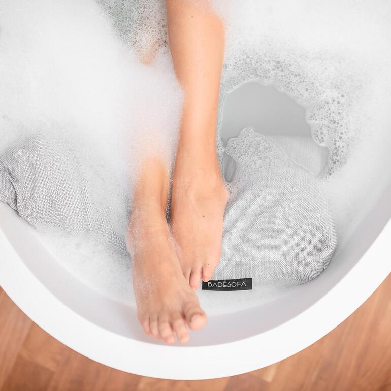 Detailaufnahme von Füßen im Schaum und Badesofa-Kissen in Badewanne.