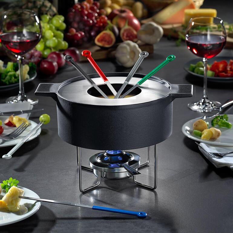 Vom feinen Metallständer sicher getragen, scheint der massive gusseiserne Topf über dem Tisch zu schweben. Mono Design-Fondue-Topf auf Tisch, umgeben von Zutaten und Wein.