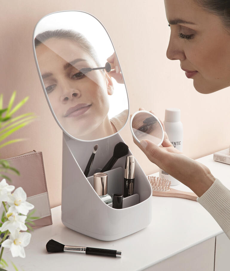 Frau schminkt sich vor einem Kosmetikspiegel mit integriertem Organizer auf einem Tisch.