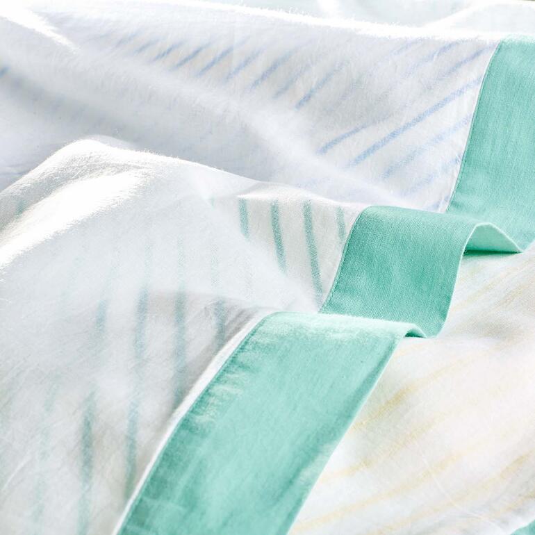 Dreilagig gefertigt mit Ober- und Unterseite aus leicht transparentem Baumwollgewebe – die Mittellage im farbigen Streifendessin schimmert effektvoll hindurch. Nahaufnahme der Breezyblanket mit einer grünen Umrandung.