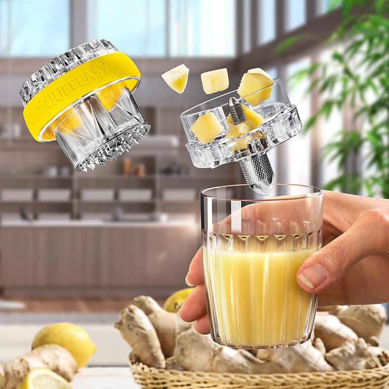 Genießen Sie Ihren Ingwer-Shot z. B. gemischt mit Orangensaft. Hand, die ein Glas mit frisch gepresstem Ingwersaft hält, während die Ingwerpresse schwebt.