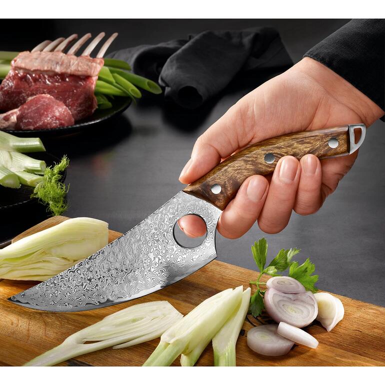 Mit Griffloch am Klingenende für sicheren Halt und präzise Führung. Damast Butcher-Messer in der Hand mit frischen Zutaten im Hintergrund.
