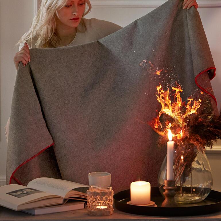 Im Notfall in Sekunden zur Hand. Das gemütliche Plaid ist zugleich eine praktische Löschdecke. Frau hält eine Feuerlöschdecke vor sich, während Flammen im Hintergrund sichtbar sind.