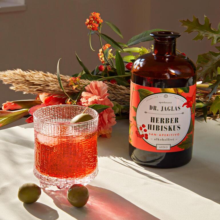 Flasche Dr. Jaglas Herber Hibiskus auf einem Tisch mit Blumen und Getränk.