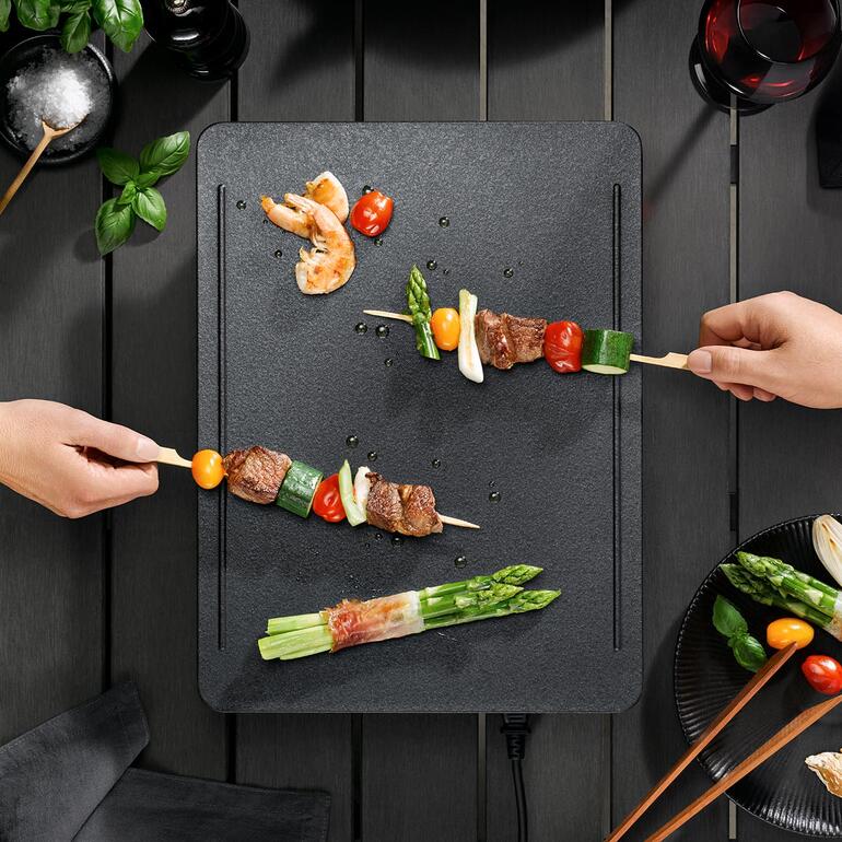Dank des innovativen, 5-schichtigen Heiz-Systems wird die Grillfläche optimal gleichmäßig beheizt. Hände stecken Spieße mit Gemüse und Fleisch auf dem Tischgrill.