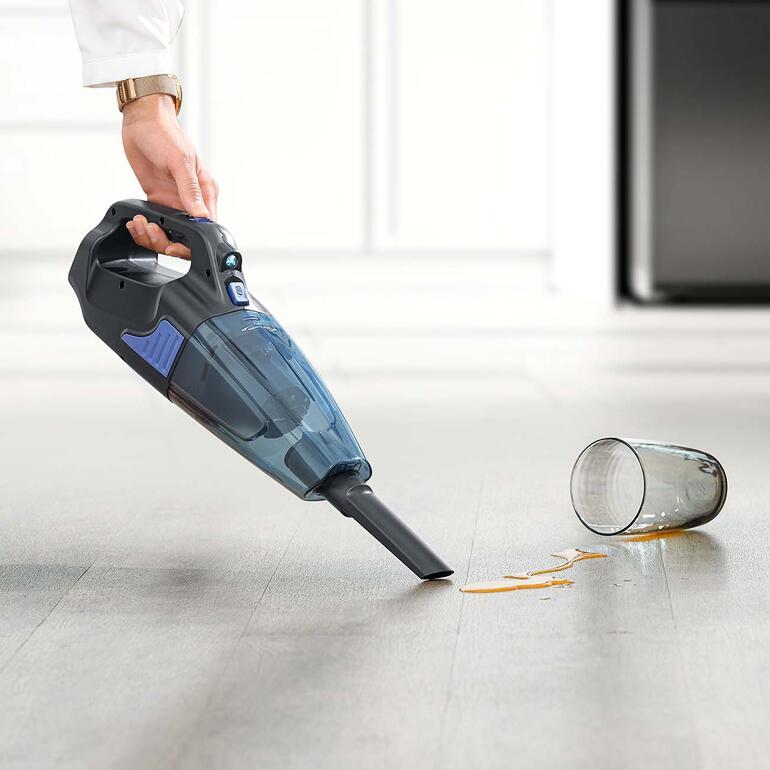 Mühelos und schnell beseitigt das Kraftpaket auch Pfützen von verschütteten Flüssigkeiten und feuchten Schmutz. Akku-Handsauger in der Hand, saugt verschütteten Saft auf einem Holzfußboden.