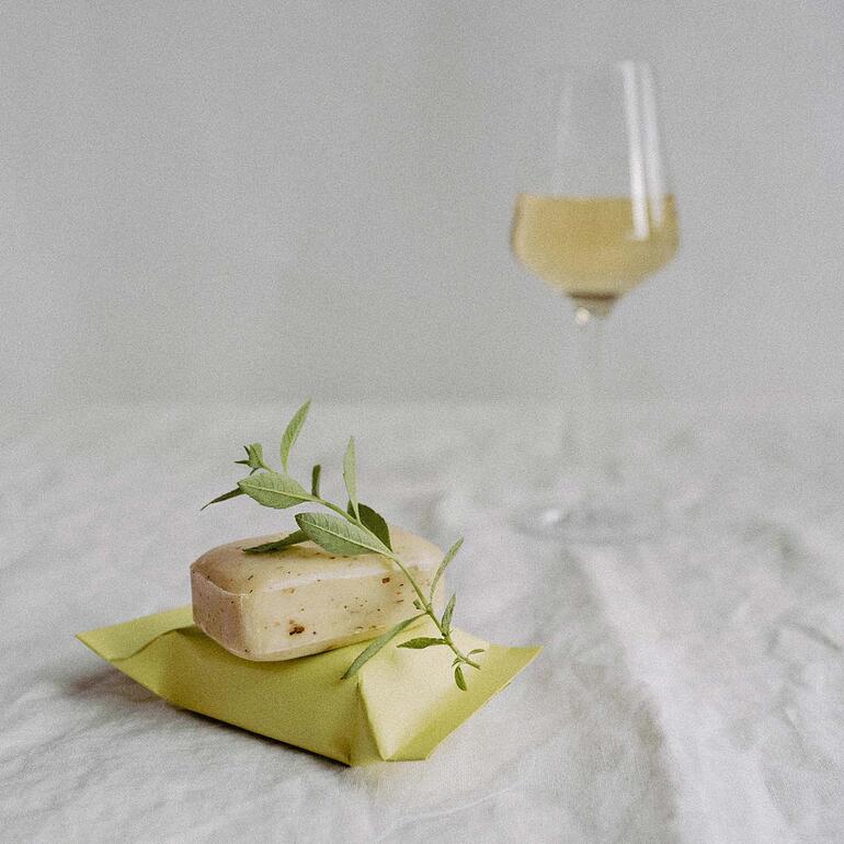 Seife The Real Wine Soap® mit Blättern dekoriert und einer Weinglas im Hintergrund.