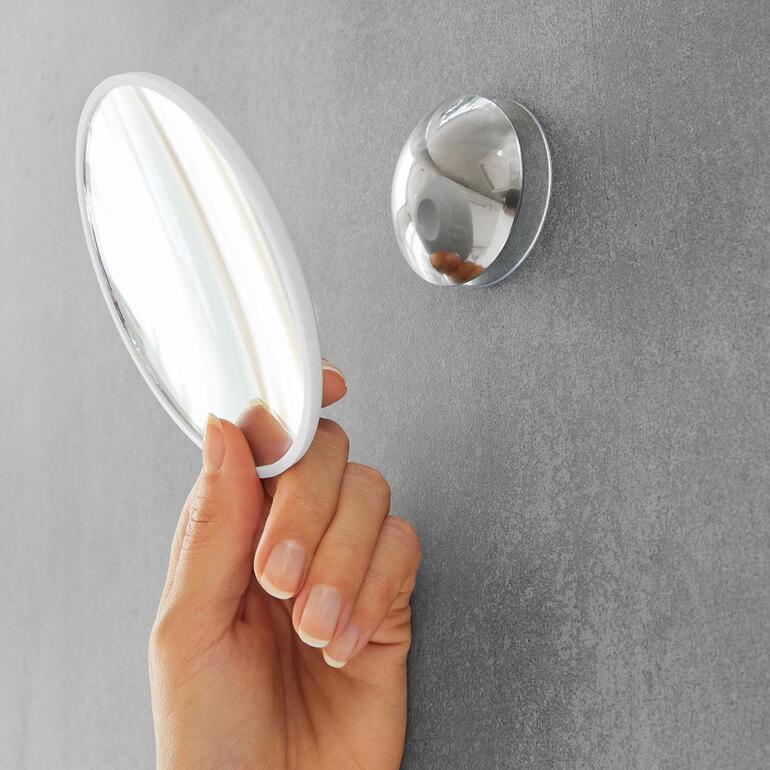 Dank Magnetkupplung einfach als Handspiegel abnehmbar. Händchen hält einen ovalen Handspiegel in der Nähe eines Wandspiegels.