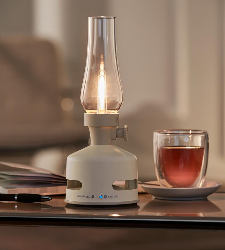 Mit integriertem Bluetooth-Lautsprecher zum Zuschalten von klangvollem Sound. LED-Öllampe in Cremefarbe auf einem Tisch mit Glas Tee.
