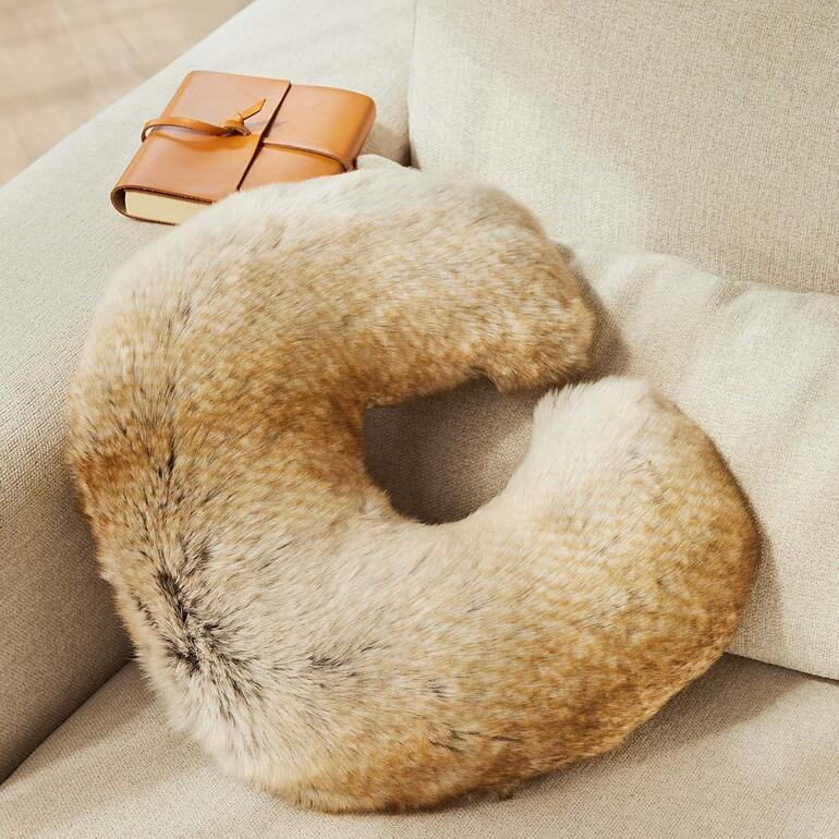 Das edel designte Nackenhörnchen ist optisch so stilvoll wie ein Dekokissen. Faux-Fur-Nackenhörnchen auf einem Sofa neben einem Buch