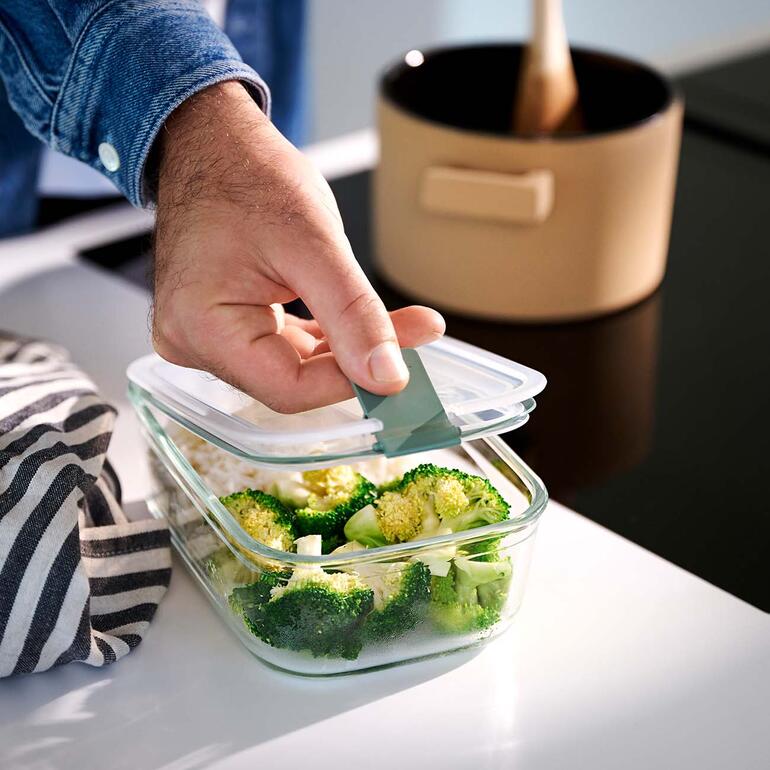 Hand, die den Deckel einer Glas-Frischhaltebox öffnet, die Brokkoli enthält.