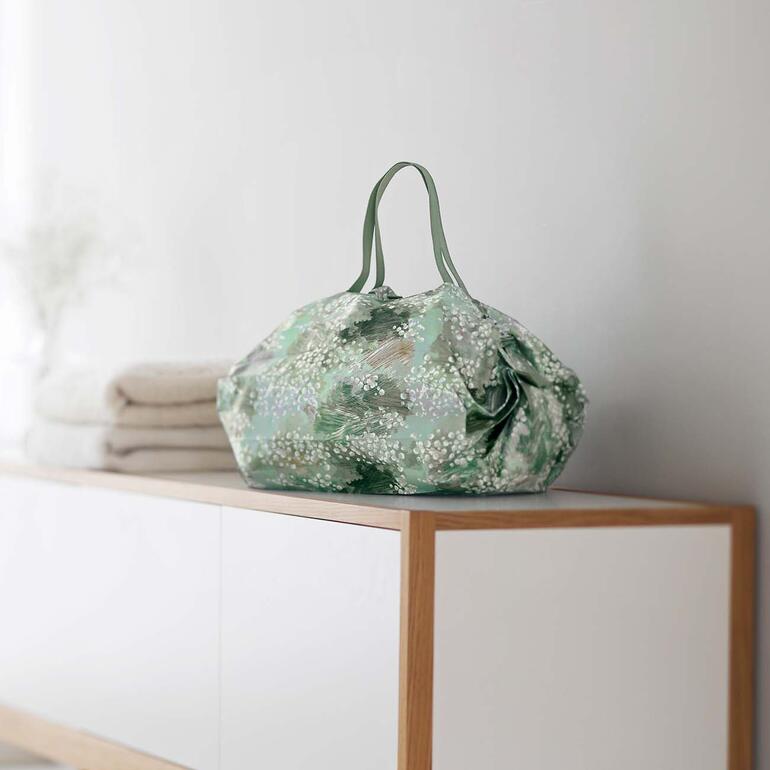 Die 15 l-Beuteltasche ist ideal für Einkauf, Reisen, Sport, Strand, … Pop-Up-Tasche auf einem Regal neben gefalteten Handtüchern.