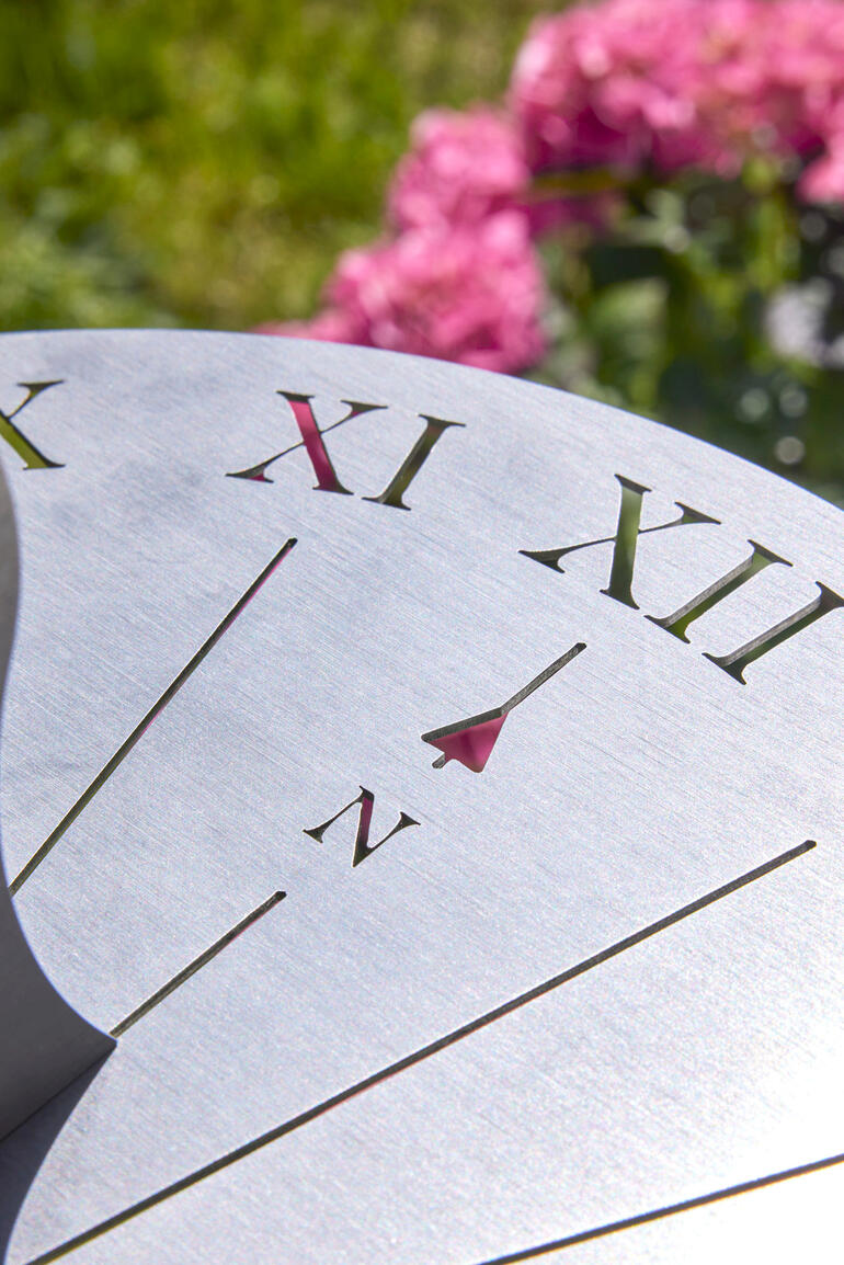 Strahlenkranz und Ziffern sind präzise per Laser in die mattierte Edelstahlscheibe gebrannt. Nahaufnahme der Ziffern auf einer Edelstahl-Sonnenuhr mit pinken Blumen im Hintergrund.