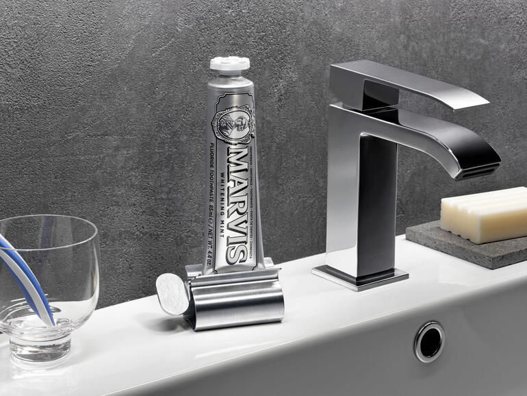 Ideal auch für Zahncreme, Ölfarbe, teure Salben, ... Edelstahl-Tubenpresse mit einer Tube Zahnpasta neben einem Wasserhahn.