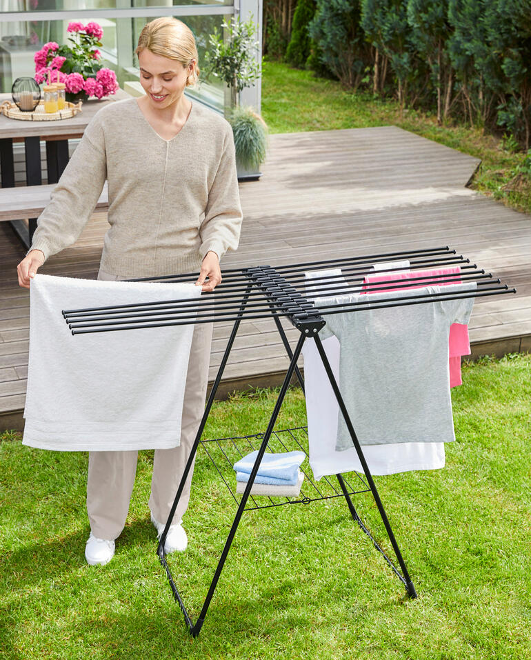Dank des rahmenlosen Designs lässt sich die Wäsche einfach von den offenen Seiten auf die Rundstäbe schieben. Frau hängt Wäsche auf einem modernen Design-Wäscheständer im Garten auf