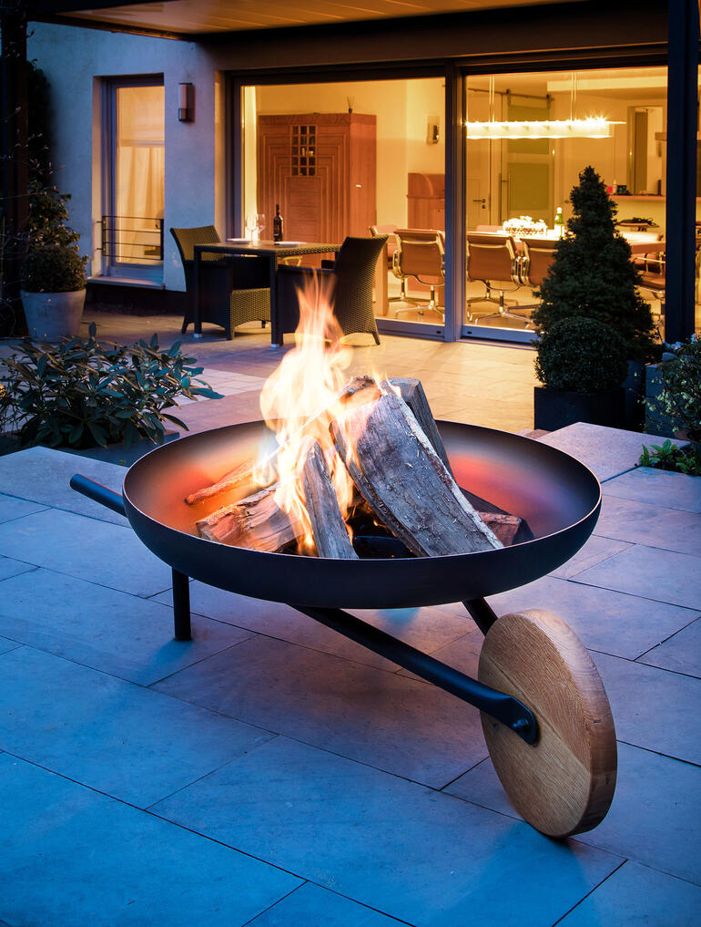 Feuerschale mit Grillfunktion, Flammen und Holzscheiten auf einer Terrasse.
