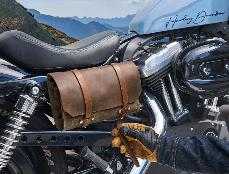 Für Ihre Motorradtouren befestigen Sie die Tasche mit den Ledergurten einfach am Gepäckträger. Echtleder-Werkzeugtasche an Motorrad befestigt, daneben eine Hand mit einem Handschuh