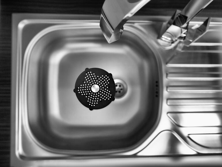 Das handtellergroße Abflusssieb ist perfekt in der Spüle, Dusche, Wanne, über dem Bodenabfluss, ... Perspektive von oben auf ein schwarzes Geruchskiller-Abflusssieb in einer Spüle.