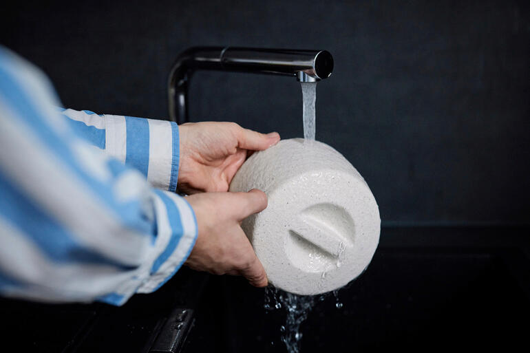 Einfach den Keramik-Butterkühler unter fließendem Wasser tränken – der Kühleffekt entsteht durch Verdunsten des aufgesogenen Wassers. Person wäscht Keramik-Butterkühler unter fließendem Wasser.