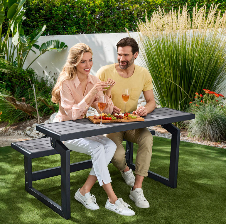 Auf Wunsch im Nu ein praktischer Essplatz. Paar sitzt an Vario-Picknicktisch im Garten und genießt Snacks und Getränke.