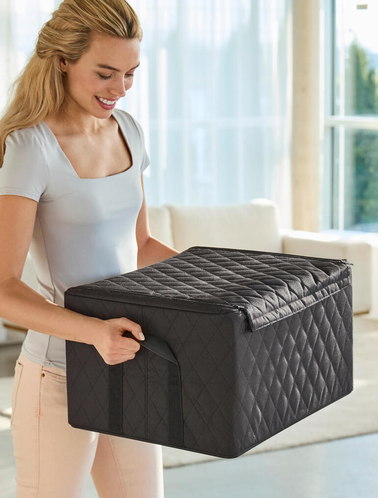 Stabile Tragegriffe ermöglichen einen komfortablen und sicheren Transport. Frau hebt eine schwarze Stapelbox mit Stepp-Rautenmuster, im Wohnzimmer.